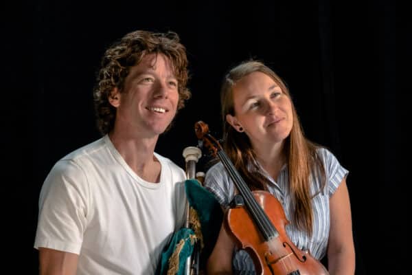 Un homme tenant une cornemuse et une femme tenant un violon sourient à quelque chose hors caméra, avec un fond sombre derrière eux.
